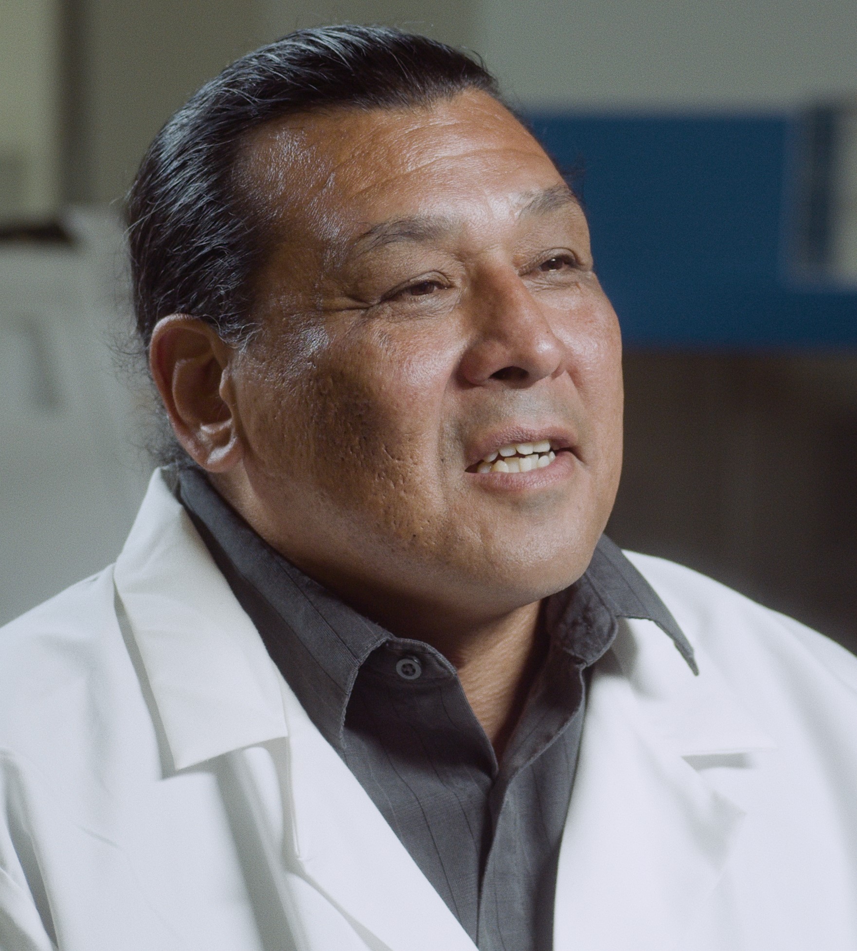Man with dark hair wearing a white lab coat and dark gray collared shirt.