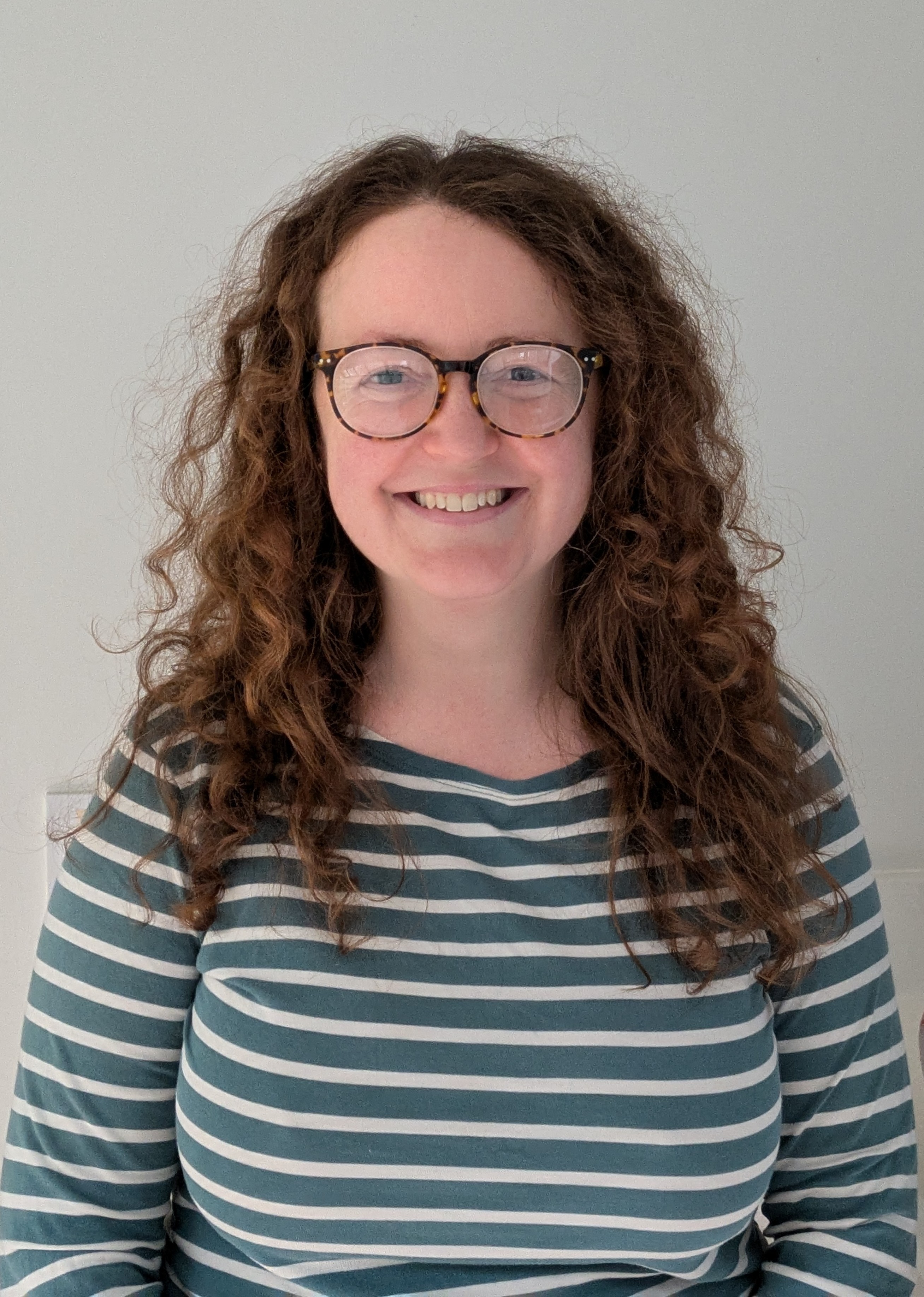 Caucasian woman smiling with long brown curly hair, glasses, and a dark green and white striped long sleeved shirt.
