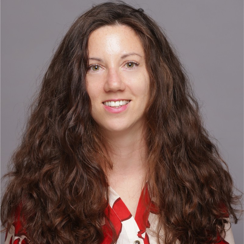 Woman smiling with long, dark brown, wavy hair, wearing a red and white blouse.
