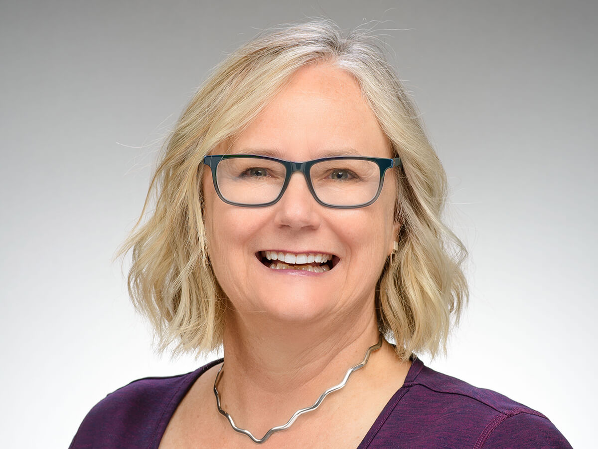 Smiling woman wearing a dark purple blouse and a necklace, with short wavy blond hair and dark-framed glasses.