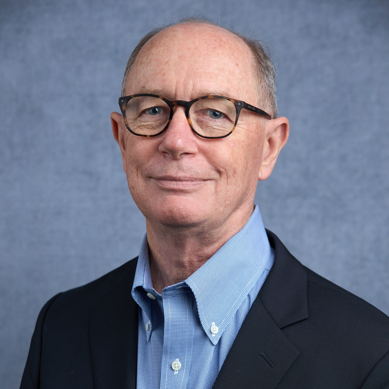 Smiling caucasian man wearing dark framed glasses, a black blazer, and a light blue collared shirt, against a denim blue backdrop.