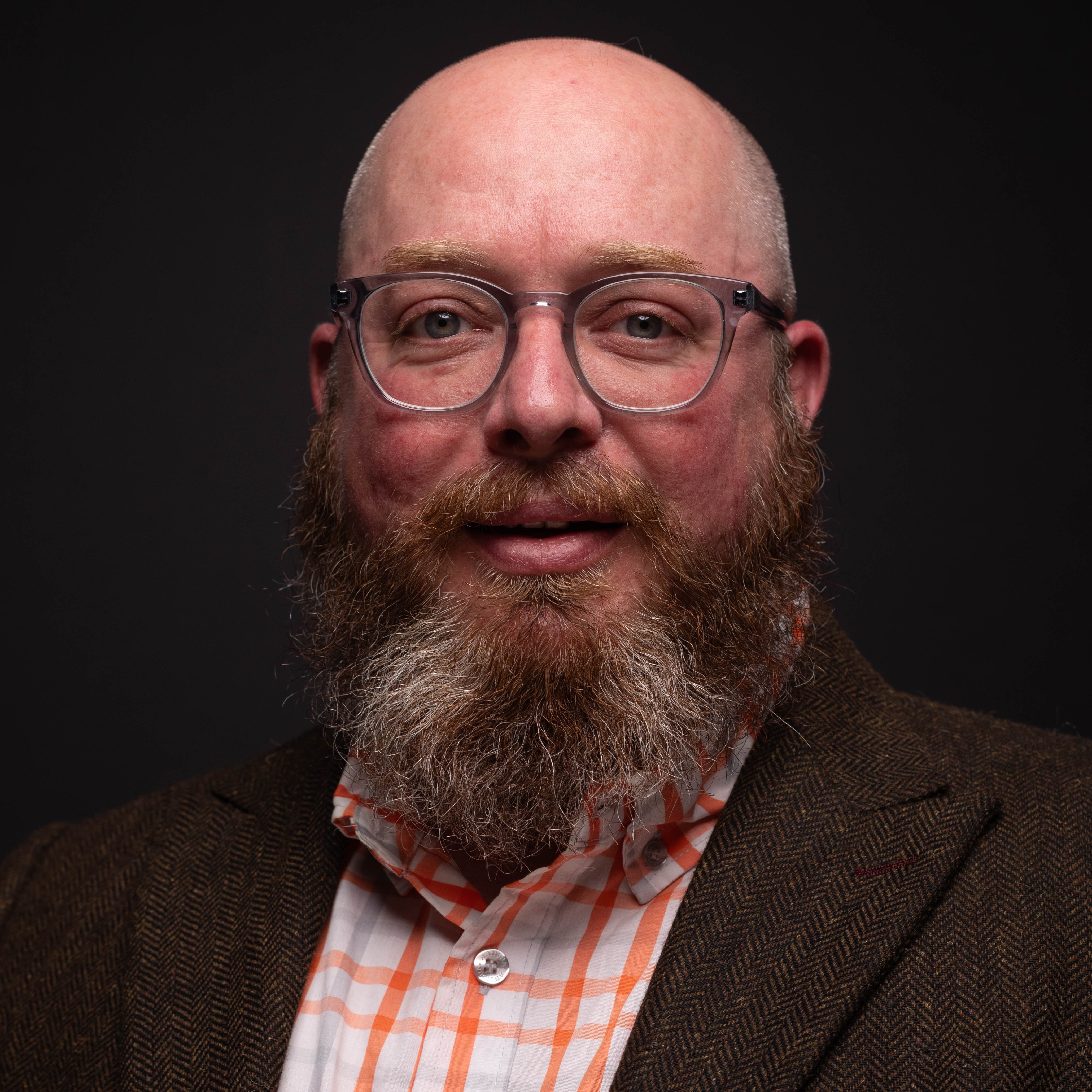 Caucasian man with a reddish-gray beard and clear glasses, wearing a brown blazer and an orange-checkered shirt, looks at the camera against a dark background.