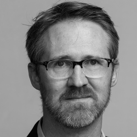 Black and white photo of a caucasian man with short hair, a neatly trimmed beard and mustache, wearing rectangular eyeglasses.