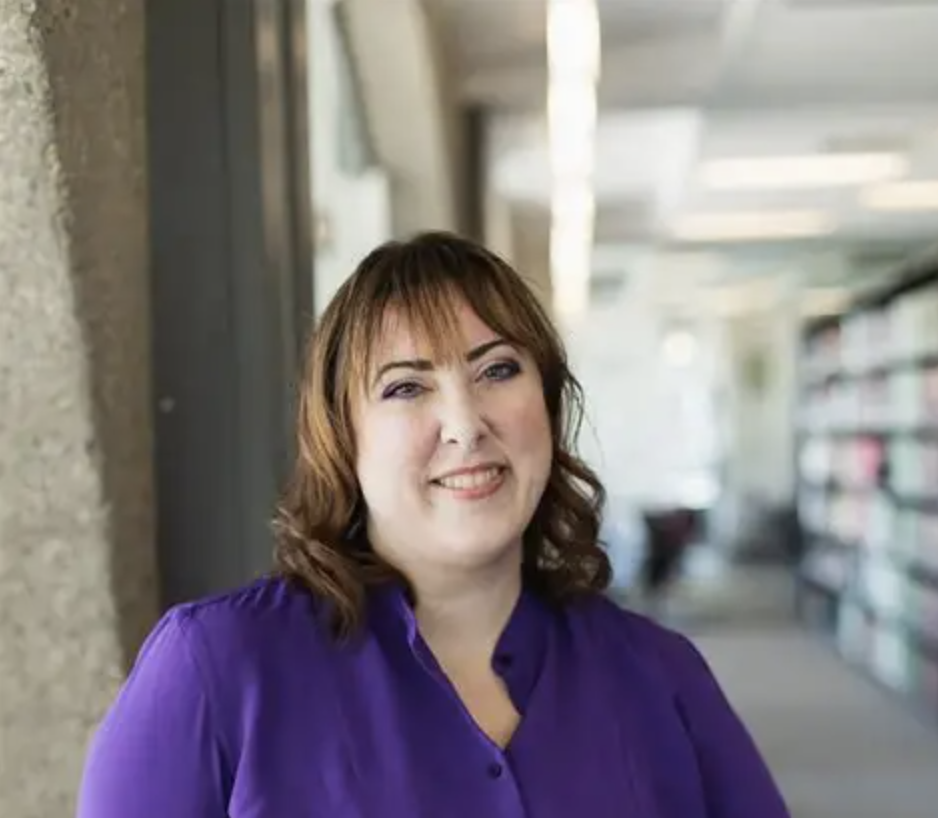 Smiling caucasian woman with shoulder-length brown hair with bangs, posing against a wall in a library.