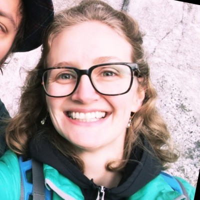 Smiling caucasian woman with large dark framed glasses, and short curly hair pulled half up.