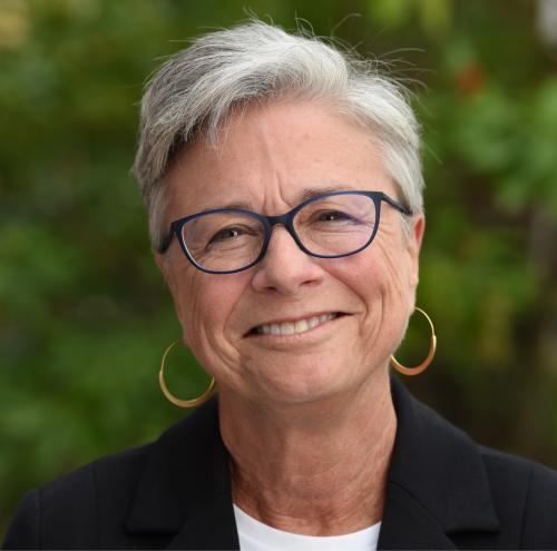 Smiling caucasian woman with short white hair, dark framed glasses, wearing gold hoop earrings and a black blazer.