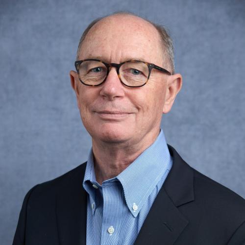Smiling caucasian man wearing dark framed glasses, a black blazer, and a light blue collared shirt, against a denim blue backdrop.
