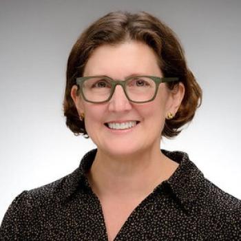 Smiling caucasian woman with short brown hair, thick olive green framed glasses, wearing a black sparkly collared shirt.